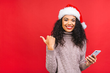 Happy excited african american woman in red santa claus hat with mobile phone isolated over red...