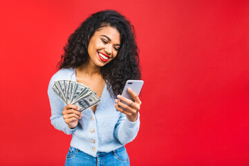Portrait of a cheerful young curly african american woman holding money banknotes and celebrating isolated over red background. Using mobile phone.