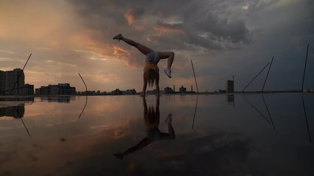 Flexible female gymnast doing handstand and calisthenic with reflection in the water on cityscape background during dramatic sunset in slow motion 