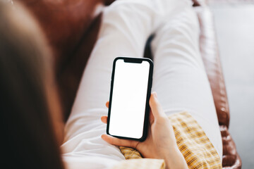 Woman holding a smartphone with a white screen mock up while sitting on the sofa at home.