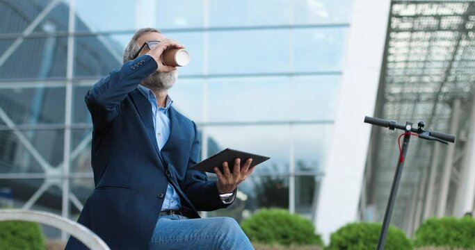 Old Caucasian man in glasses sitting outdoor at electric scooter, sipping drink and using taplet device. Male drinking coffee at street. Grandfather browsing online. Businessman pensioner.