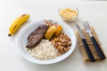 Plate of rice, beans, steak and cassava. With cutlery and manioc flour and banana for dessert. White wooden background in Brazilian kitchen copy space, top view. Food concept. Pasta concept.