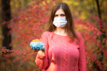 A girl in a medical mask with a pumpkin in a pink sweater against the background of an autumn...