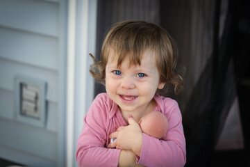 Cute Adorable 18 Month Old Baby Girl with Big Blue Eyes and Brown Curly Hair playing with Doll. Happy healthy kid play alone on the Porch.