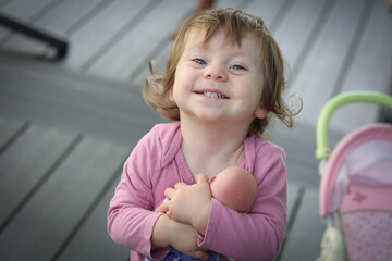 Cute Adorable 18 Month Old Baby Girl with Big Blue Eyes and Brown Curly Hair playing with Doll. Happy healthy kid play alone on the Porch.