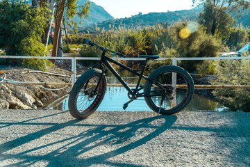 Fototapeta premium A black new bike leaning in the middle of a small river bridge on a sunny day with lens flare. The shadow is falling o the road. Behind nature reflecting in water. Old pipes lying at shore .