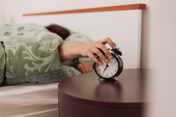 Caucasian woman in bed reaching out for analogue alarm clock to turn it off at 7 am, close up shot.