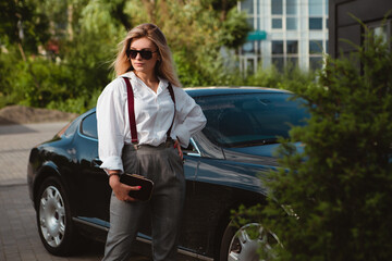 Portrait of young business boss woman standing near expensive luxury car with clutch bag, dressed in white shirt, trousers, suspenders, looking away. Image with copy space.