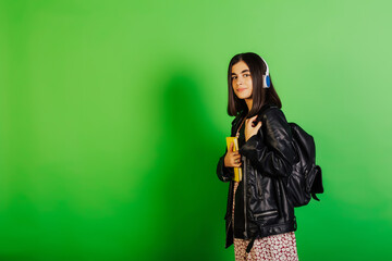 Charming student in dress, leather black jacket and black backpack are holding yellow notebook and listening music with wireless headphones. Copy space.