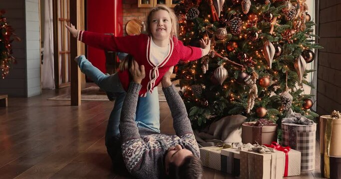 Strong dad lying on wooden floor at decorated house hall on Christmas Eve play with small daughter lifts up child stretched arms imitates plane, imagines flying in air. Family travel, xmas fun concept