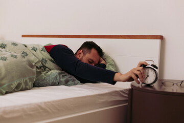 Fototapeta premium Sleepy man in bed reaching out for analogue alarm clock to turn it off at 7 am