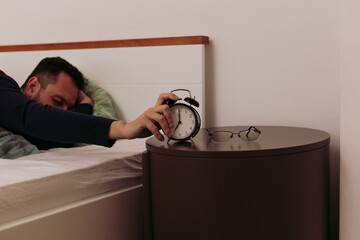 Sleepy  man in bed reaching out for analogue alarm clock to turn it off at 7 am