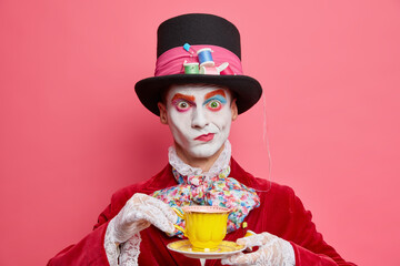 Horizontal shot of serious male hatter poses with cup of tea wears hat has manners of aristocratic gentleman dresses for masquerade carnival poses indoor. Halloween and entertainment concept - obrazy, fototapety, plakaty