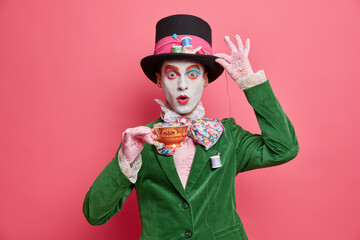Photo of shocked male has image of mysterious hatter from wonderland wears bright makeup poses with cup of tea dressed in aristocratic clothes isolated on pink background. Halloween party concept - obrazy, fototapety, plakaty