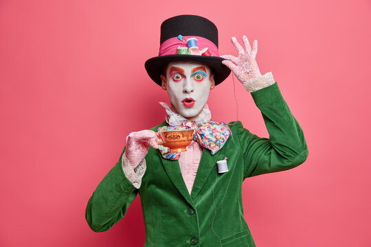 Photo of shocked male has image of mysterious hatter from wonderland wears bright makeup poses with cup of tea dressed in aristocratic clothes isolated on pink background. Halloween party concept