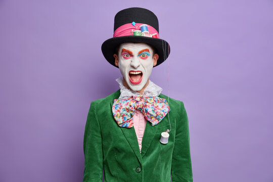 Emotional hatter screams loudly dressed in festive costume poses against purple background on halloween party has insane look. Crazy joker exclaims from negative emotions keeps mouth widely opened