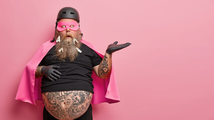 Surprised plump man with thick beard dressed like superhero raises palm and holds invisible object demonstrates copy space has big belly sticking out from undersized black t shirt. Costumed guy © WHstudio Leushin N