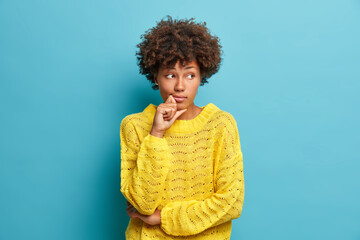 Serious pensive woman with Afro hair looks away and stands in thoughtful pose thinks about problems and difficulties dressed in warm yellow sweater isolated on blue background makes decision
