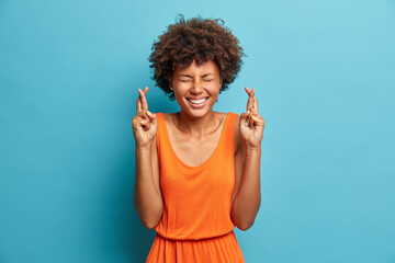 Superstitious cheerful African American woman keeps fingers crossed wishes for good luck believes in dreams come true laughs with closed eyes dressed in orange summer dress isolated on blue background