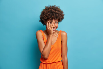 Happy shy woman makes face palm looks through fingers dressed in orange dress poses against blue...
