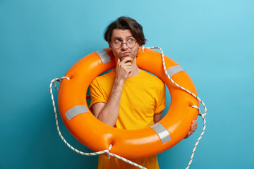 Photo of thoughtful male lifesaver poses with inflated swim ring holds chin and looks away...