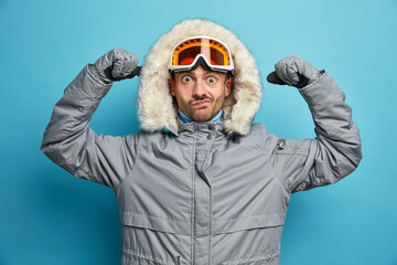 Winter time concept. Surprised male skier raises hands and shows his strength has active rest in mountains goes snowboarding wears outerwear looks seriously at camera isolated on blue background