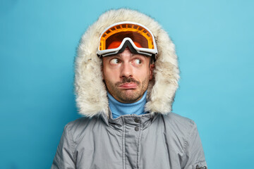 Close up shot of man hiker looks thoughtfully aside wears ski goggles and winter jacket with hood has active rest during vacation enjoys favorite sport isolated on blue background. Skier in mountains