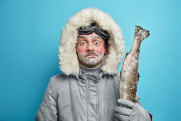 Puzzled surprised frozen man spends long hours outdoor during severe cold day dressed in grey winter jacket and gloves holds fish wears ski glasses isolated on blue background. Frosty weather - Powered by Adobe