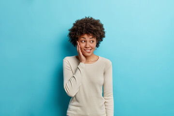 Fototapeta na wymiar Studio shot of lovely thoughtful young African American woman bites lips and looks away with pensive expression thinks about way out wears long sleeved casual jumper isolated on blue background