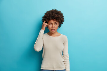 Fototapeta na wymiar Hesitant unaware woman with curly hair rubs temple and has confused expression tries to remember something dressed in casual clothes isolated on blue background. Doubtful dark skinned female