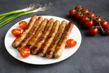 Grilled, roasted, delicious sausages on a white plate on a gray concrete background with green onion and cherry tomatos.