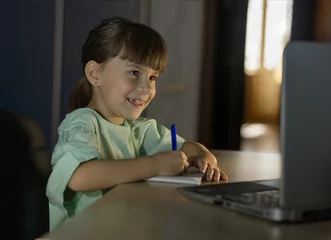 Photo sur Plexiglas Doux monstres Enseignement à distance. Une fille heureuse apprend l& 39 anglais en ligne avec un ordinateur portable à la maison. La petite fille regarde l& 39 ordinateur portable