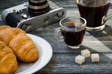 Still life with cup of coffee and croissant on the wooden background. Old retro camera and postcard are near the cup.