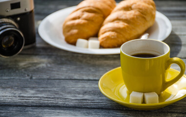 Still life with cup of coffee and croissant on the wooden background. Old retro camera and postcard are near the cup.