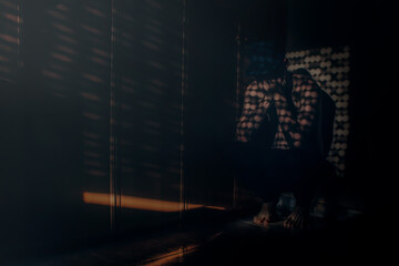 A squatting person hidden in the shadows with his hands covering his face is illuminated by rays of light that filter through the blind of the bedroom window.