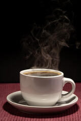Fototapeta premium Coffee steaming in a cup with saucer isolated on a black background on a red surface 