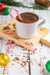 Hot chocolate in a white cup with red and white candy canes Red, green and gold Christmas balls. Worn white wood background. Brown kitchen cabinet. Christmas background. Breakfast.