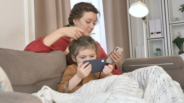 Positive young mother strokes head of little girl holding mobile phones sitting under knitted plaid on sofa in room closeup