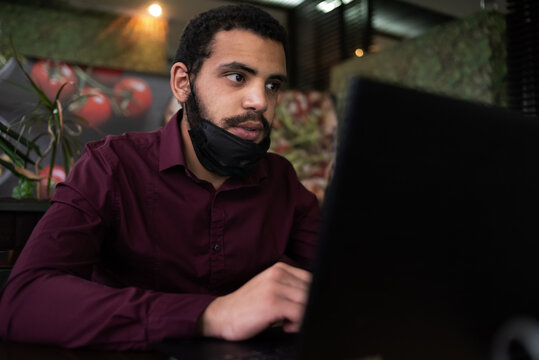 African businessman or developer wearing surgical face mask in the restaurant and typing on laptop - concept on new normal everyday lifestyle and people at work in covid-19 pandemic season.