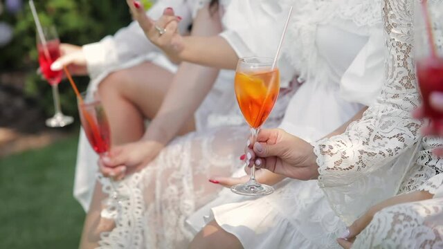 Girls in white dresses sit side by side and hold champagne and cocktails glasses in their hands, close up.