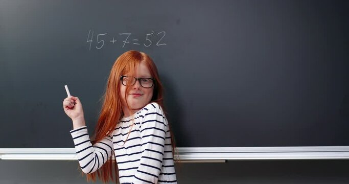 Rear on Caucasian red-haired girl standing at blackboard and writing equations with chalk. Schooling concept. Back view on schoolgirl doing math in school and turning happy to camera. Mathematics.