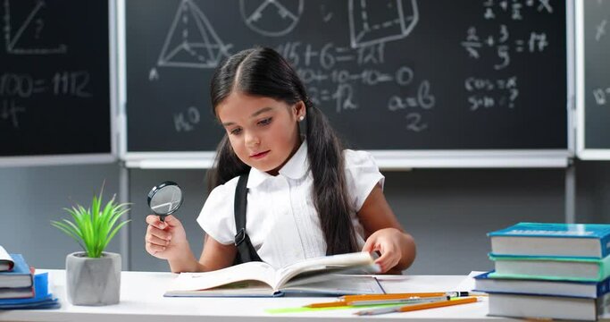 Cute little teen girl smiling and reading textbook with magnifying glass at desk in classroom. Pretty small schoolgirl flipping pages of book and looking at pictures. School concept.