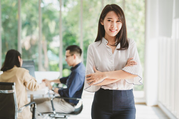 Obraz na płótnie Canvas Portrait of beautiful young Asian businesswoman standing arms crossed with colleague in background in meeting room in office