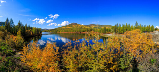 Panoramic shot. A beautiful flat surface of the lake against the background of colored hills and the blue sky. Fascinating view of the forest lake