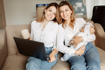 Portrait of happy three generation, middle aged woman with her adult daughter sit at the sofa, young mother working from home, caring grandmother babysit with newborn baby girl