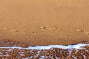 Footprints of bare human feet walking from left to right along smooth yellow sand along the seashore with transparent water with a narrow strip of white sea foam