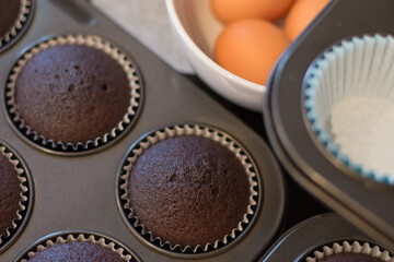 Close up of chocolate cupcakes in baking trays with a bowl of eggs in the background