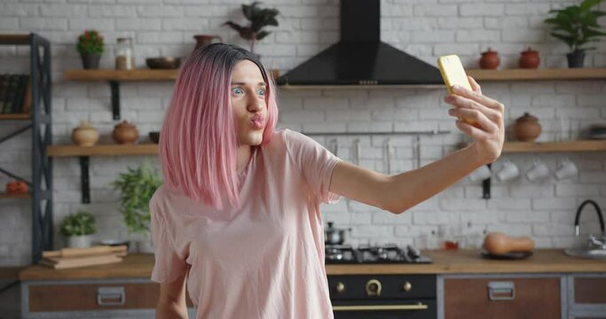 Pretty young trans woman with stylish pink wig makes selfie with contemporary smartphone posing in decorated kitchen at home