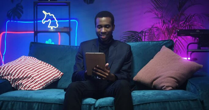 Smiled cheerful good looking stylish African American man in black clothes sitting on couch. Handsome guy using tablet computer on sofa in living room with neon lights. Male tapping, typing and