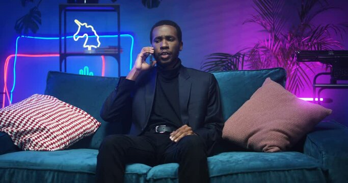 Good looking young African American man calling on cellphone in room with neon lights. Handsome black stylish guy talking on phone while sitting on couch with LED light. Male speaking on mobile on